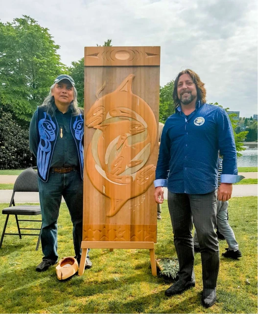 Two men standing beside a large indigenous wood carving display during an outdoor cultural event.